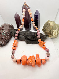 A pink and red coral coral bead necklace with chunky coral beads and small round garnet beads,rhodonite and pink opal and chocolate rhodocrosite  displayed on a white surface with crystal points and rocks in the background.