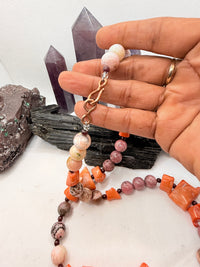 A pink and red coral coral bead necklace with chunky coral beads and small round garnet beads,rhodonite and pink opal and chocolate rhodocrosite  displayed on a white surface with crystal points and rocks in the background.