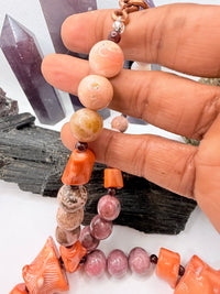A pink and red coral coral bead necklace with chunky coral beads and small round garnet beads,rhodonite and pink opal and chocolate rhodocrosite  displayed on a white surface with crystal points and rocks in the background.