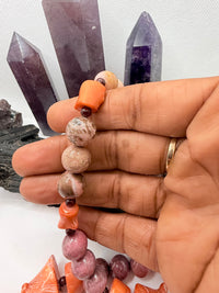 A pink and red coral coral bead necklace with chunky coral beads and small round garnet beads,rhodonite and pink opal and chocolate rhodocrosite  displayed on a white surface with crystal points and rocks in the background.