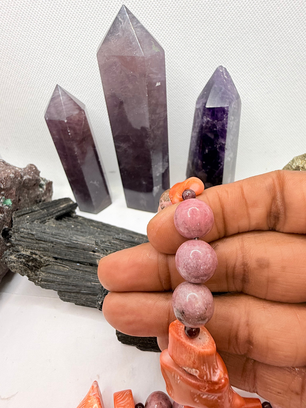 A pink and red coral coral bead necklace with chunky coral beads and small round garnet beads,rhodonite and pink opal and chocolate rhodocrosite  displayed on a white surface with crystal points and rocks in the background.