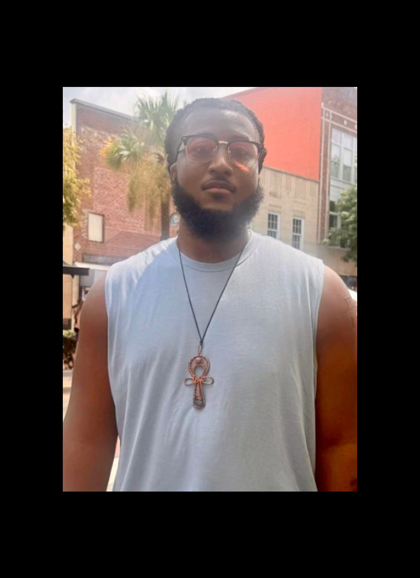 A man wearing a light blue sleeveless t-shirt and a black cord necklace with a copper-colored Ankh pendant, standing outdoors in front of brick buildings.