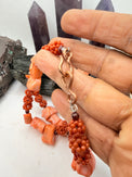 A pink and red coral coral bead necklace with chunky coral beads and small round garnet beads, displayed on a white surface with crystal points and rocks in the background.