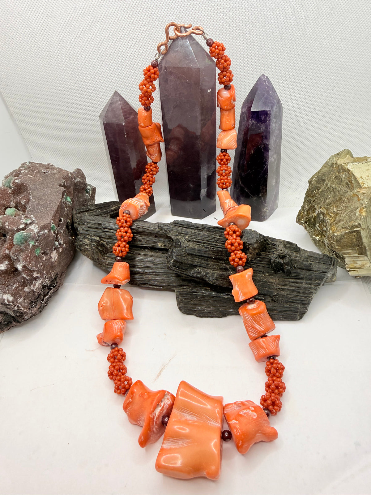 A pink and red coral coral bead necklace with chunky coral beads and small round garnet beads, displayed on a white surface with crystal points and rocks in the background.