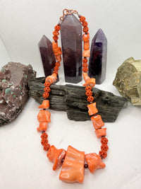 A pink and red coral coral bead necklace with chunky coral beads and small round garnet beads, displayed on a white surface with crystal points and rocks in the background.